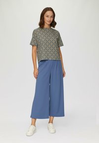 Gemustertes kurzärmliges Oberteil in Dunkelblau, Creme und Orange mit geradem Schnitt. Kombiniert mit hellblauen weit geschnittenen Hosen und weißen Sneakers.
