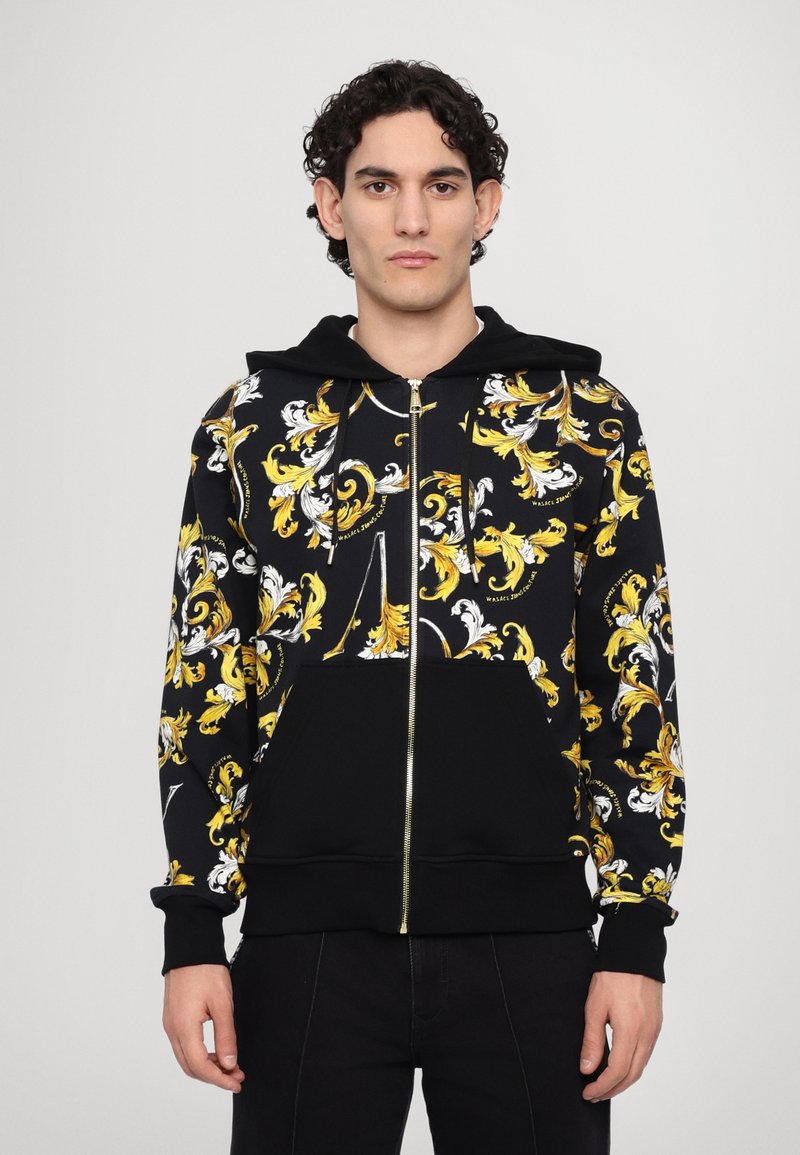 Μαύρη ζακέτα με φερμουάρ, με έντονο κίτρινο και λευκό μπαρόκ μοτίβο, μαλακό ύφασμα, μπροστινές τσέπες και κουκούλα με κορδόνι.