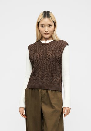 Junge Frau mit langen blonden Haaren trägt ein weißes, geripptes Langarmshirt unter einer braunen, strukturierten, gestrickten ärmellosen Weste und lockeren braunen Hosen.