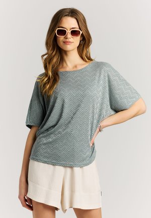 Mujer con blusa gris con estampado suelta, pantalones cortos crema, gafas de sol cuadradas beige y pulseras plateadas, posando con una mano en la cadera.