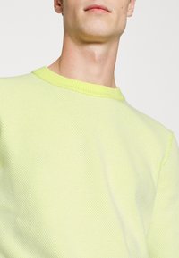 Limonkowo zielony sweter z dzianiny o fakturowanym wzorze, okrągłym dekoltem i ściągaczami na rękawach. Krótkie rękawy i lekki materiał.