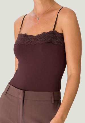 Kobieta ubrana w ciemnobrązową koszulkę na cienkich ramiączkach z koronkową lamówką, zestawioną z brązowymi, wysokim stanem spodniami na neutralnym tle.