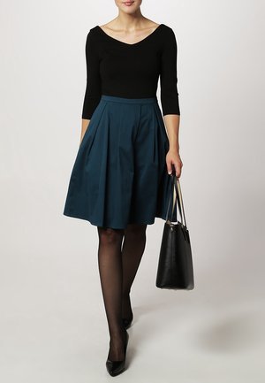 Schwarzes Longsleeve-Oberteil mit rundem Ausschnitt, kombiniert mit einem türkisfarbenen A-Linien-Rock. Das Modell trägt schwarze, durchsichtige Strumpfhosen und hält eine schwarze Handtasche.