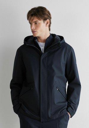 Junger Mann trägt eine marineblaue Kapuzenjacke mit seitlichen Reißverschlusstaschen, die Hände in den Taschen, blickt nach links vor einer schlichten hellen Wand.