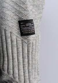 Harmaa teksturoitu neulekangas, jossa on ribbikuvio ja musta etiketti, jossa lukee "Petrol Ind" ja "1973", esittelee perinnetietoista suunnittelua.