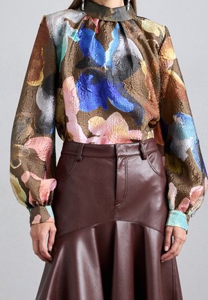 Femme portant un chemisier à manches longues à fleurs multicolores avec un col montant et une jupe en cuir marron à ourlet évasé.