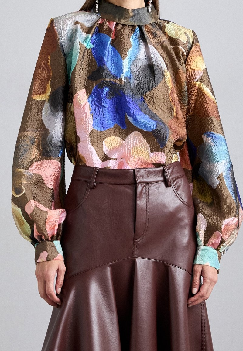 Femme portant un chemisier à manches longues à fleurs multicolores avec un col montant et une jupe en cuir marron à ourlet évasé.
