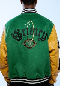 Chaqueta letterman verde y amarilla con el texto bordado "Grimey" y un gráfico, que presenta un emblema negro y dorado, cuello y puños acanalados.