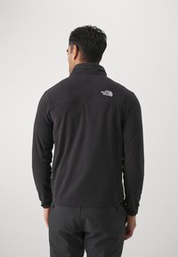 Mężczyzna w czarnej kurtce polarowej i ciemnych spodniach, odwrócony plecami, z logo The North Face widocznym na górnej części pleców.