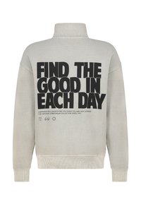 Grijze sweatshirt met hoge kraag en een gedurfde zwarte tekst: "VIND HET GOEDE IN IEDERE DAG." Inclusief extra kleinere tekst en grafische patronen.