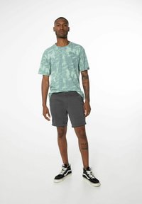 T-shirt verde menta a tie-dye con maniche corte, abbinata a pantaloni corti grigi. Il modello indossa scarpe da ginnastica nere alte. Tatuaggio sulla gamba visibile.