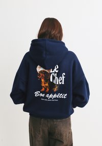 Námořnická modrá oversized mikina s grafikou, na které je jezevčík, text "Le Chef Bon appétit" a kulatý kuchařský klobouk jako akcent. Měkká textura.