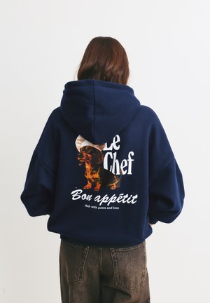 Temno modra oversize kapuca s grafiko, ki prikazuje osliča, besedilom "Le Chef Bon appétit" in okroglim kuhinjskim klobukom kot poudarkom. Mehek material.