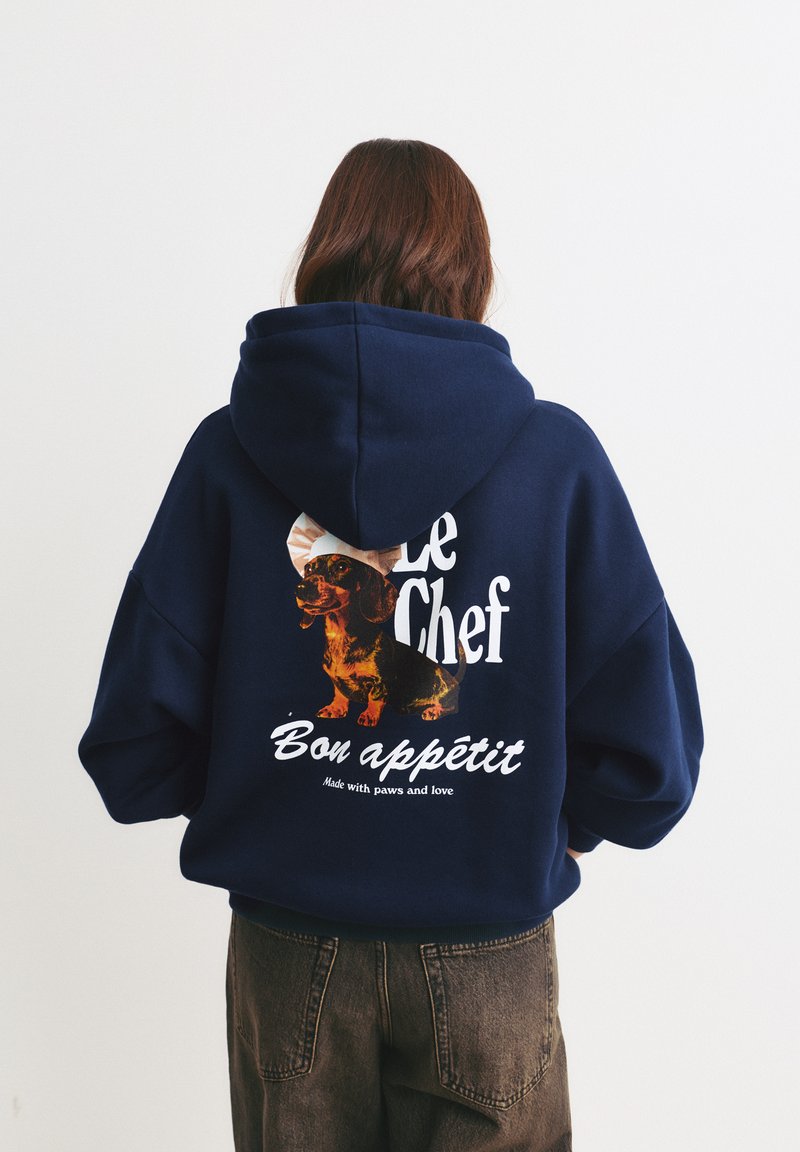 Felpa oversize blu navy con una grafica che rappresenta un bassotto, la scritta "Le Chef Bon appétit" e un cappello da cuoco rotondo come accento. Texture morbida.