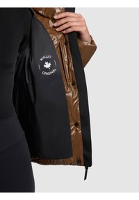 Mano che tiene aperta una giacca marrone e nera mostrando la fodera interna nera con il logo "Khujo Original" e una tasca laterale verticale con cerniera.