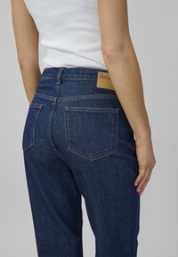 Osoba v tmavomodrých denimových nohaviciach a bielom topánkach bez rukávov, zobrazená zozadu na jednoduchom šedom pozadí.