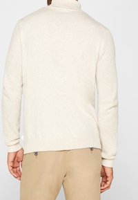 Pull en tricot beige clair avec un col montant, un corps texturé et des poignets côtelés. Associé à un pantalon marron clair avec des boutons décoratifs.