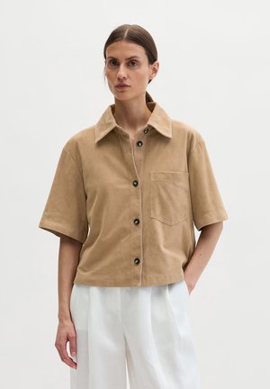 Frau mit braunen Haaren im niedrigen Pferdeschwanz, trägt ein kurzärmeliges, beigefarbenes Hemd mit Knöpfen und weiße plissierte Hosen, steht vor einem einfarbigen Hintergrund.