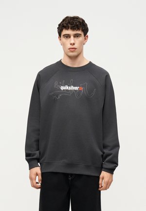 Jeune homme aux cheveux courts et bouclés portant un sweat Quiksilver gris foncé et un pantalon noir, debout devant un fond clair uni.