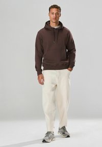 Brauner Kapuzenpullover mit einer Vorderasche, kombiniert mit lockeren, cremefarbenen Hosen und grauen Sportschuhen. Einfarbiger Hintergrund.