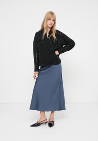 Mulher loira de pé com a mão na cintura, vestindo uma blusa preta de mangas longas, uma saia midi azul e sapatos tipo slingback pretos com bico fino.