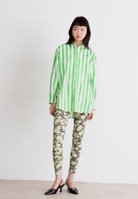 Grønn og hvit stripete knapp-up skjorte, parret med blomsterprydede leggings. Modellen har på seg svarte høye hæler. Minimalistisk bakgrunn.
