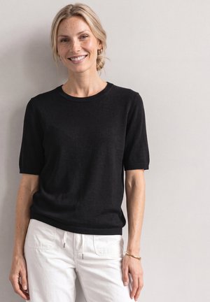 Femme souriante aux cheveux blonds attachés en un chignon bas, vêtue d'un haut noir à manches courtes et d'un pantalon blanc, debout devant un mur clair uni.