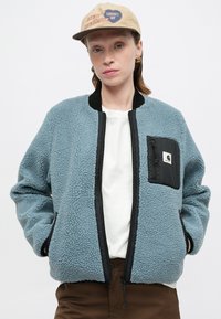 Világoskék bolyhos bomberdzseki fekete díszítéssel és egy zsebbel. Fehér ing felett viselve; barna nadrággal és bézs sapkával párosítva.