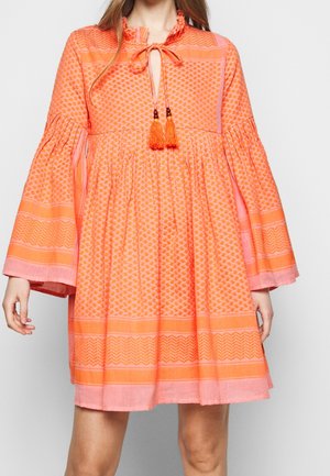 Mujer vistiendo un vestido naranja suelto hasta la rodilla, con mangas largas acampanadas y cordones con borlas en un escote fruncido.