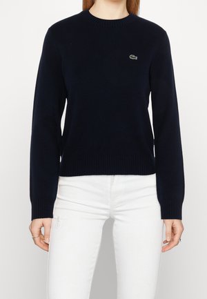 Kobieta w granatowym, długim rękawie z okrągłym dekoltem, z małym logo krokodyla, ubrana w obcisłe białe spodnie na jednolitym tle.