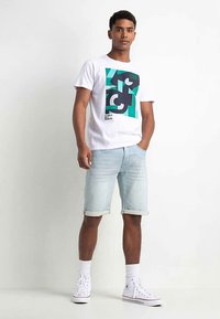 Weißes T-Shirt mit einem geometrischen, schwarz-türkisfarbenen grafischen Motiv auf der Vorderseite, kombiniert mit hellblauen Jeansshorts, die am Saum umgeschlagen sind.
