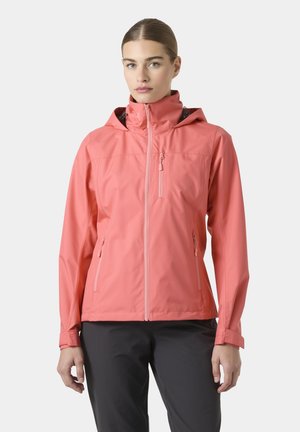 Mujer con chaqueta impermeable con capucha color coral y pantalones negros, de pie frente a un fondo liso gris claro.