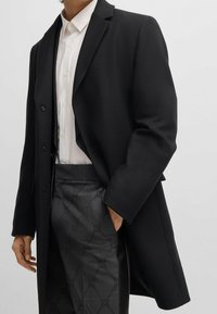 Cappotto lungo nero in misto lana, con colletto a punta e chiusura a bottoni. Sotto, una camicia bianca è abbinata a pantaloni a quadri.