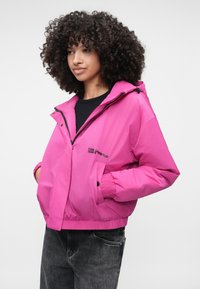 Jachetă izolată roz cu glugă, manșete elastice și două buzunare laterale. Prezintă un logo vizibil pe față și un design confortabil.