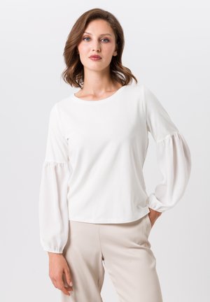 Vrouw met schouderlang haar, gekleed in een witte blouse met lange mouwen en geplooide mouwen, en lichtbeige broek, staand met een hand in de zak.