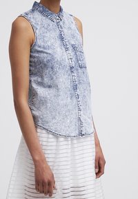 Chemise en denim délavé sans manches avec poche avant, associée à une jupe blanche semi-transparente à rayures, portée par une personne debout.