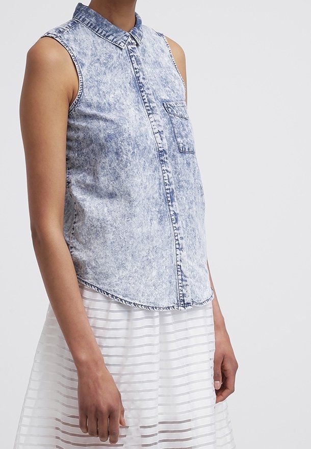 Chemise en denim délavé sans manches avec poche avant, associée à une jupe blanche semi-transparente à rayures, portée par une personne debout.