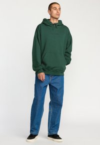 Sudadera verde con cordón, gran bolsillo frontal y logotipo bordado. Combinada con vaqueros anchos azules y zapatillas negras.