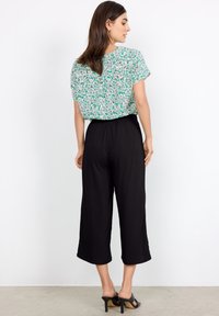 Blusa de estampado floral en verde y blanco con mangas cortas, combinada con pantalones negros recortados y sandalias de tacón negro.