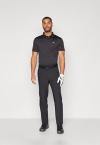 Polo nero con colletto e logo, abbinato a pantaloni neri. Una cintura nera e un guanto da golf bianco completano l'outfit.
