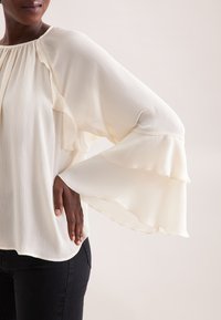 Blusa color crema con maniche lunghe e volants, caratterizzata da una texture plissè e un collo rotondo, abbinata a pantaloni neri aderenti.