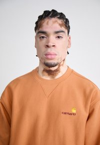 Orange sweatshirt med en rund udskæring og ribstrikkede manchetter, forsynet med et lille gult og burgunder broderet logo på brystet.