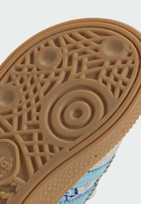 Semelle d'une chaussure en caoutchouc gomme brun clair avec des motifs circulaires et en grille sur la bande de roulement, et une partie de la tige bleue à motifs visible.