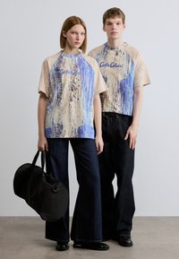 Γραφικά t-shirts σε μπεζ χρώμα με σχέδιο πιτσιλιές μπλε μπογιάς και κείμενο "Carlos Clucci". Σκούρα τζιν καμπάνα. Μαύρη στρογγυλή τσάντα duffel.