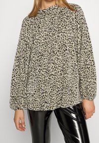 Blusa beige chiaro con stampa leopardata nera, texture plissettata, scollo alto e maniche lunghe a palloncino, abbinata a pantaloni neri lucidi.