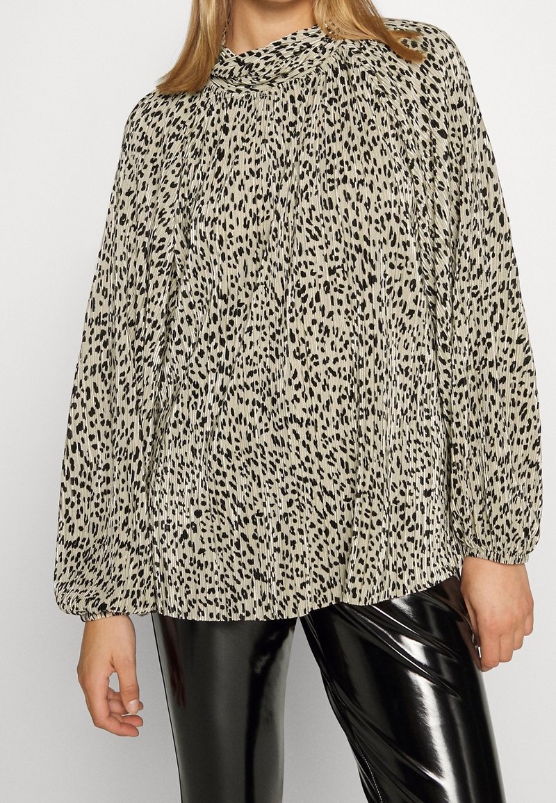 Blusa beige chiaro con stampa leopardata nera, texture plissettata, scollo alto e maniche lunghe a palloncino, abbinata a pantaloni neri lucidi.