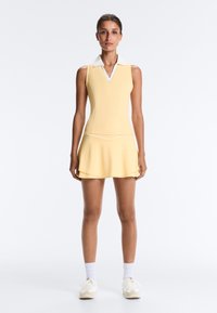 Femme debout portant un haut sans manches jaune avec col et une jupe assortie, des chaussettes blanches et des baskets blanches sur un fond uni.
