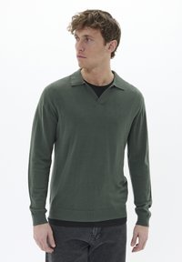 Pulover verde cu mâneci lungi, cu decolteu în V �și guler pliat. Țesătura pare moale și ușoară, purtat peste un tricou negru, asociat cu blugi închiși la culoare.