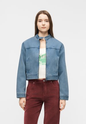 Tânără cu părul lung și brunet, purtând o jachetă scurtă din denim albastru, un tricou alb cu imprimeu și pantaloni roșii din catifea cu talie înaltă.