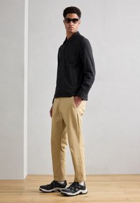 Casaco cinza escuro de botões com colarinho e bolso frontal, combinado com calças bege ajustadas e ténis pretos com detalhes brancos.